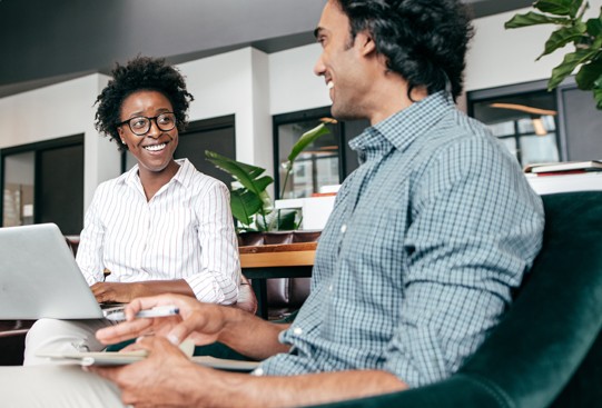 A woman with curly hair and glasses smiles while sitting with a laptop. A man with wavy hair responds positively. They are in a modern, plant-filled workspace.