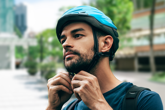 A man is fastening the chin strap of a blue bicycle helmet. He appears focused and is outdoors, surrounded by greenery and urban buildings in the background.