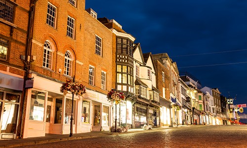 Rows of charming, historical buildings line a cobblestone street, illuminated by warm lights, under a darkening sky, conveying a peaceful evening atmosphere in an inviting town.