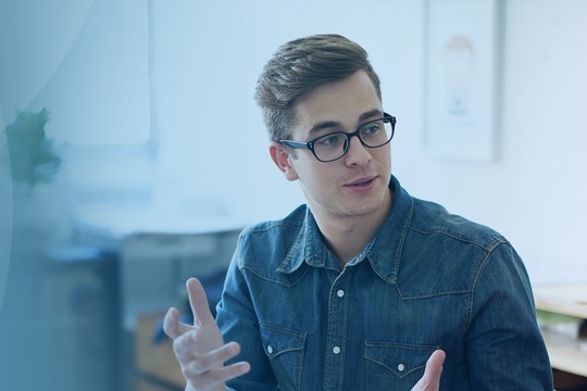 A man in glasses gestures while speaking to a woman, both engaged in a conversation. They are in a bright, modern office environment with desks and plants visible in the background.