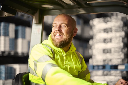 A man in a bright yellow hoodie smiles while operating a forklift in a warehouse filled with stacked goods, conveying a sense of positivity and engagement in his work environment.