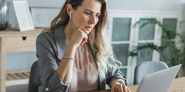 A woman wearing earphones sits at a wooden table, focused on her laptop. She rests her chin on her hand, with a light-filled, minimalist workspace and plants in the background.