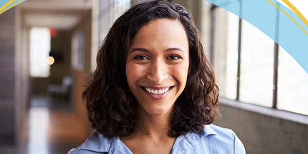 A woman with curly hair smiles warmly while wearing a light blue shirt. She stands in a well-lit hallway featuring large windows, enhancing the inviting atmosphere.