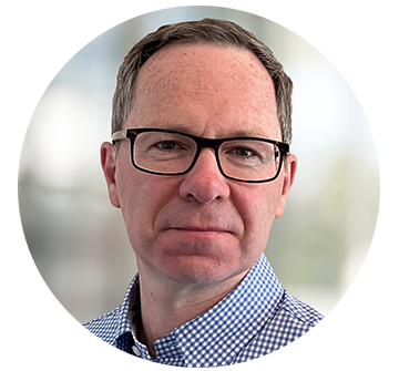 A man with glasses and short hair is facing the camera, wearing a checkered blue shirt, against a blurred light background that suggests an indoor environment.