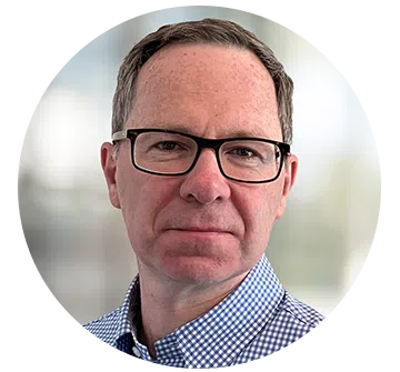 A man with glasses and short hair is facing the camera, wearing a checkered blue shirt, against a blurred light background that suggests an indoor environment.