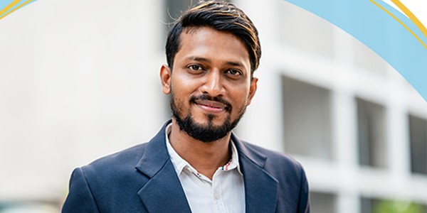 A man with a beard and light hair stands outdoors, smiling confidently in a dark blazer over a light shirt, with modern buildings visible in the background.