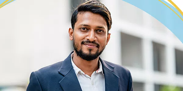 A man with a beard and light hair stands outdoors, smiling confidently in a dark blazer over a light shirt, with modern buildings visible in the background.