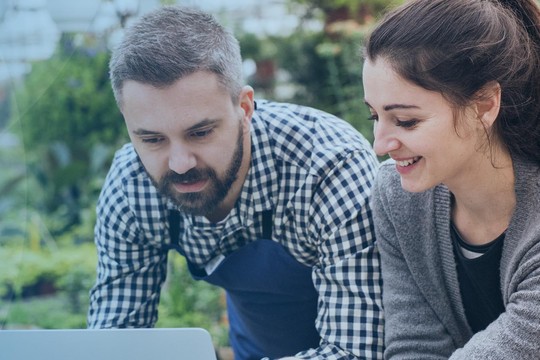 Two individuals, a man and a woman, are collaborating on a laptop, smiling and engaged. They are situated in a lush, green environment with plants around them, suggesting a serene outdoor setting.