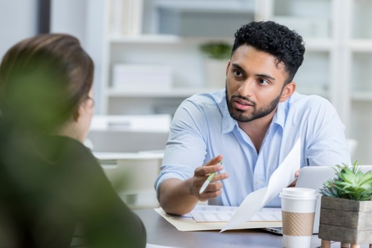 A man in a light blue shirt is discussing documents while gesturing with a pen. He sits at a table with a coffee cup and potted plants in a modern office setting.