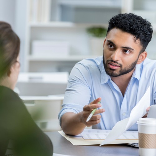 A man in a light blue shirt is discussing documents while gesturing with a pen. He sits at a table with a coffee cup and potted plants in a modern office setting.
