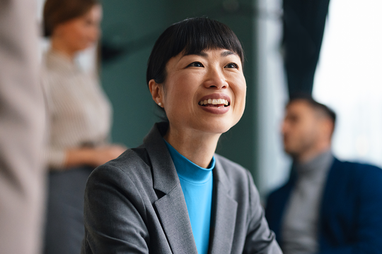 A woman in a gray suit smiles warmly while seated at a table. She is engaged in conversation, with two individuals partially visible in the background, suggesting a professional setting.