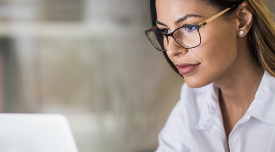 A woman with glasses is focused on her laptop, typing. The setting is a bright, modern workspace with blurred shelves visible in the background, suggesting a productive environment.