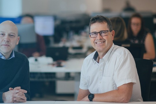 Two men are sitting at a desk, smiling and engaging with the camera in a modern office environment. In the background, colleagues work at computers, suggesting a collaborative atmosphere.