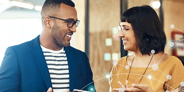 A man and woman stand close together, smiling and engaging in conversation. They hold a smartphone, surrounded by a modern, bright workspace featuring wooden textures and soft lighting.