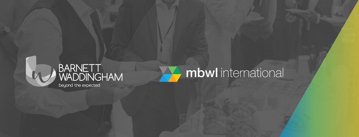 Individuals are holding plates with food at a gathering, with a backdrop featuring logos and branding elements for Barnett Waddingham and mbwl international, suggesting a business event.