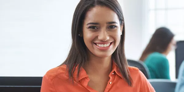 A woman with long brown hair smiles while seated at a desk. In the background, blurred figures are working at computers, suggesting a professional office environment.