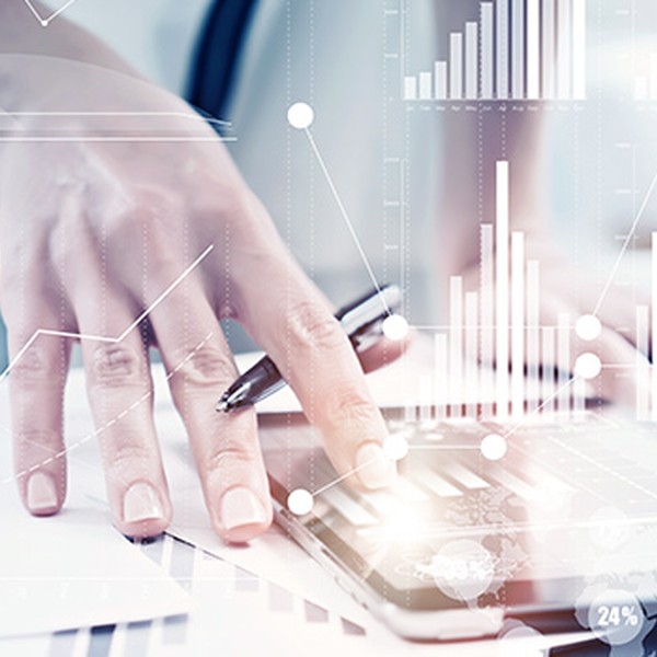A hand holds a pen over a calculator while other papers and digital graphs are visible. The context suggests a busy office environment focused on financial analysis or data management.