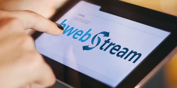 A hand with a finger touches a tablet screen displaying the logo "hwebstream," featuring blue text with arrows indicating motion. The background is softly blurred, suggesting a warm indoor setting.