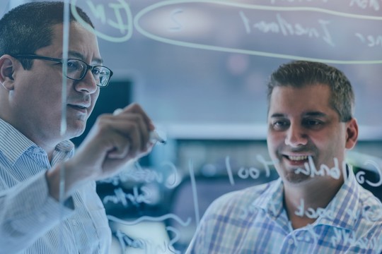 Two men collaborate in a modern office setting, one writing on a glass board while the other observes and smiles, engaged in a discussion about ideas or concepts.