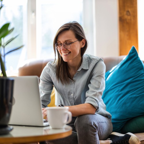 A woman sits comfortably on a sofa, smiling while typing on a laptop. She is surrounded by colorful cushions and a potted plant, with soft natural light illuminating the room.