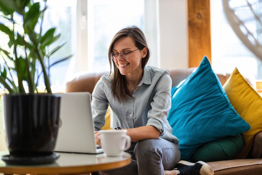 A woman sits comfortably on a sofa, smiling while typing on a laptop. She is surrounded by colorful cushions and a potted plant, with soft natural light illuminating the room.