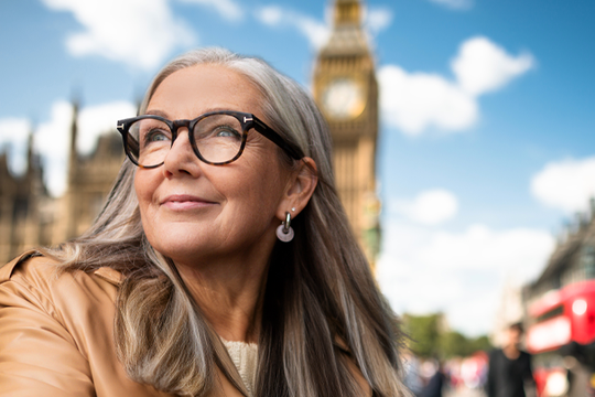 A woman with long gray hair and glasses gazes upward, smiling softly. In the background, the iconic Big Ben clock tower is visible, under a bright blue sky with fluffy clouds.