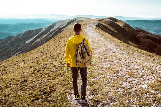 A person in a yellow jacket walks along a rocky path on a grassy hillside. The background features rolling mountains under a clear sky, suggesting a scenic outdoor adventure.