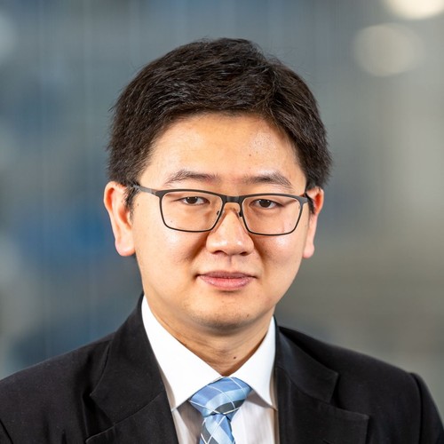 A man wearing glasses and a suit with a tie is facing the camera. The background is blurred, suggesting an indoor professional environment.