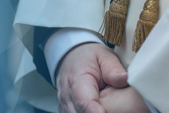 A close-up view of a person's hands, clasped together, wearing a white robe with ornate detailing and golden tassels. The background features a soft blue gradient.