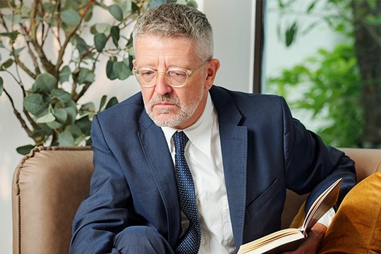 A middle-aged man in a suit sits on a couch, reading a book with a serious expression. Lush greenery is visible through a window in the background.