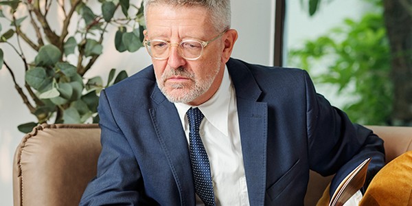 A middle-aged man in a suit sits on a couch, reading a book with a serious expression. Lush greenery is visible through a window in the background.