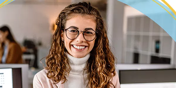 A young woman with curly hair and glasses smiles confidently. She stands in an office space, suggesting a professional atmosphere, while colleagues are visible in the background.