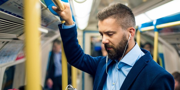 A man in a blue blazer stands on a subway train, holding a pole with one hand while using his phone with the other. He listens to music with earphones.