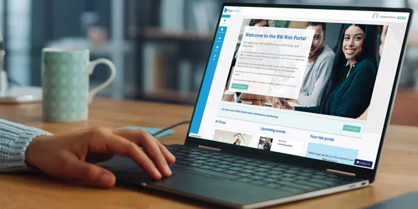 A hand rests on a laptop keyboard, displaying a webpage titled "Welcome to the BW Risk Portal!" Nearby, a light blue mug is visible on a wooden table in a cozy indoor setting.