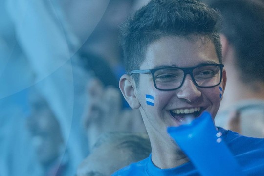 A group of enthusiastic fans at a sporting event are cheering and smiling, displaying joy and excitement in a vibrant atmosphere filled with other supporters and decorations in shades of blue.