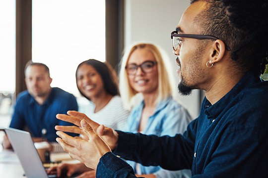 A man with glasses gestures expressively while speaking to three smiling individuals at a modern conference table, engaged in conversation. Natural light filters through large windows.