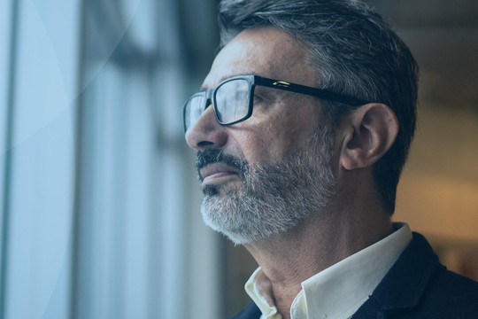 A man with glasses stands by a window, gazing thoughtfully outside. The interior is modern, with soft lighting, and tall city buildings are visible in the background.