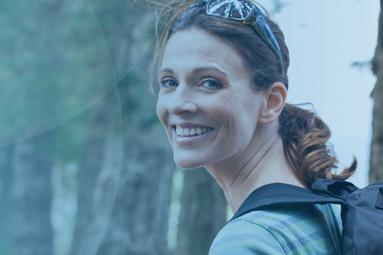 A woman smiles while walking through a forest, wearing a backpack and sunglasses. She is positioned near tall trees, suggesting an outdoor or hiking context.
