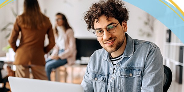 A young man with curly hair and glasses is smiling while working on a laptop. Two people are conversing in the background, suggesting a collaborative office environment.