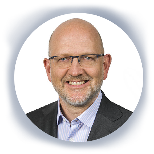 A middle-aged man with glasses and a beard smiles, wearing a dark blazer over a light blue collared shirt, set against a plain white background.