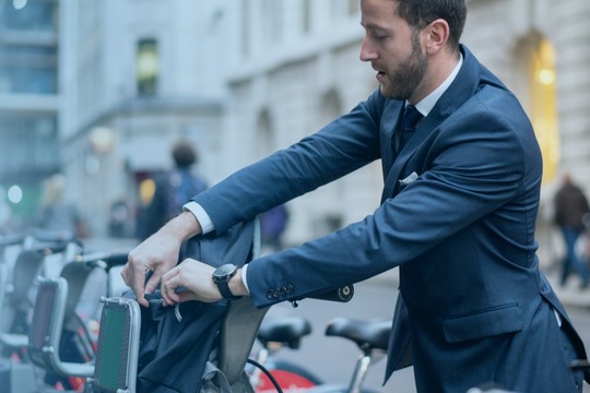 A man in a suit is unlocking a bicycle at a bike-sharing station on a city street, surrounded by other parked bicycles and a blurred urban backdrop.