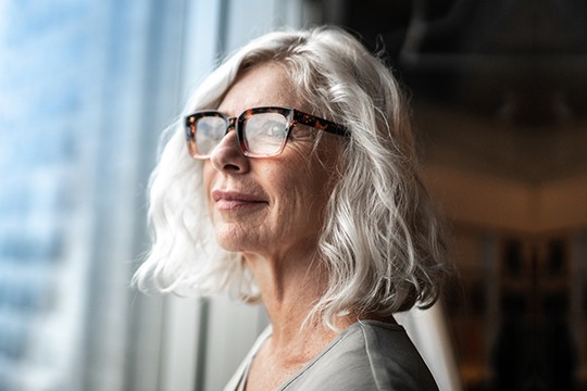 A woman with shoulder-length, wavy gray hair wears stylish glasses, gazing thoughtfully out a large window, with a blurred indoor setting in soft focus behind her.