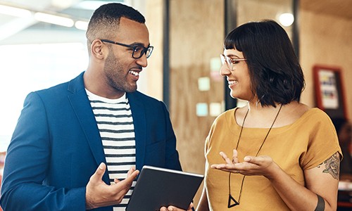 A man and a woman are engaged in conversation, with the man holding a tablet, in a modern workspace with wooden accents and soft lighting.