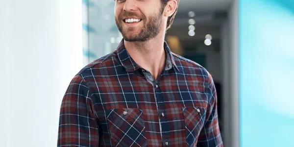 A man wearing a plaid shirt is holding a tablet and smiling. He stands in a modern, well-lit environment with blue accents and blurred background elements.