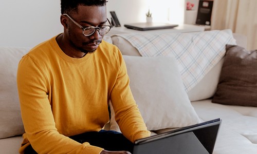 A man in a yellow sweater types on a laptop while sitting on a light-colored sofa. The room is cozy with soft pillows and a blanket draped nearby.