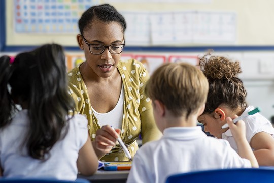 A teacher engages with young students at a table, guiding their drawing activities. The classroom is colorful, with educational posters and charts in the background. Children focus on their tasks.