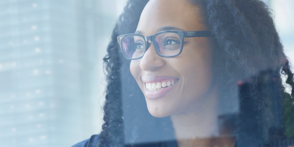 A woman with curly hair and glasses smiles while looking out a large window. Urban buildings are visible in the blurred background, suggesting a bright, modern city environment.