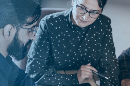 A group of three individuals collaborates closely, reviewing documents and discussing ideas in a modern office setting, with a soft blue background enhancing the professional atmosphere.