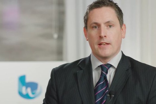 A man in a dark suit and striped tie speaks directly, conveying information. Behind him, a logo on a white backdrop hints at a business context, with a professional, modern setting.