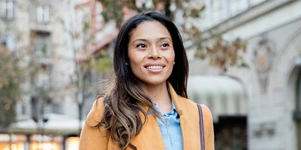 A woman with long hair, wearing a yellow coat over a blue shirt, smiles while walking in a city street lined with buildings and trees, suggesting a pleasant atmosphere.